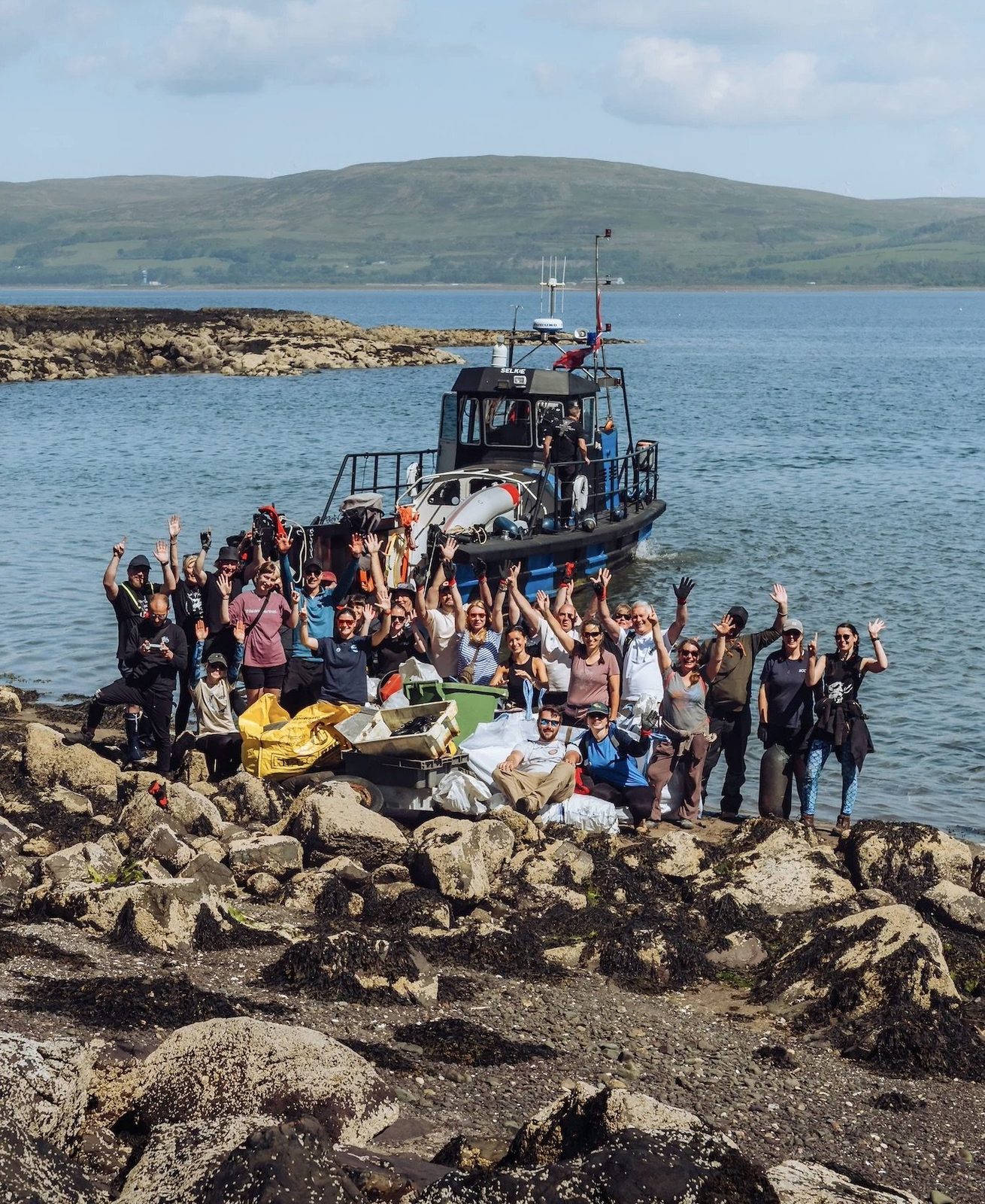 A large group on a rocky shoreline beside a boat during a Project One Wave clean-up.